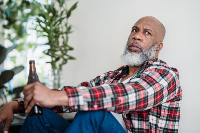 Bearded man in plaid shirt, sitting and holding a beer indoors.