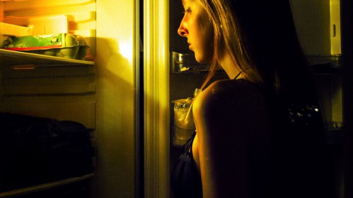 Woman standing at an open fridge late at night looking for a snack